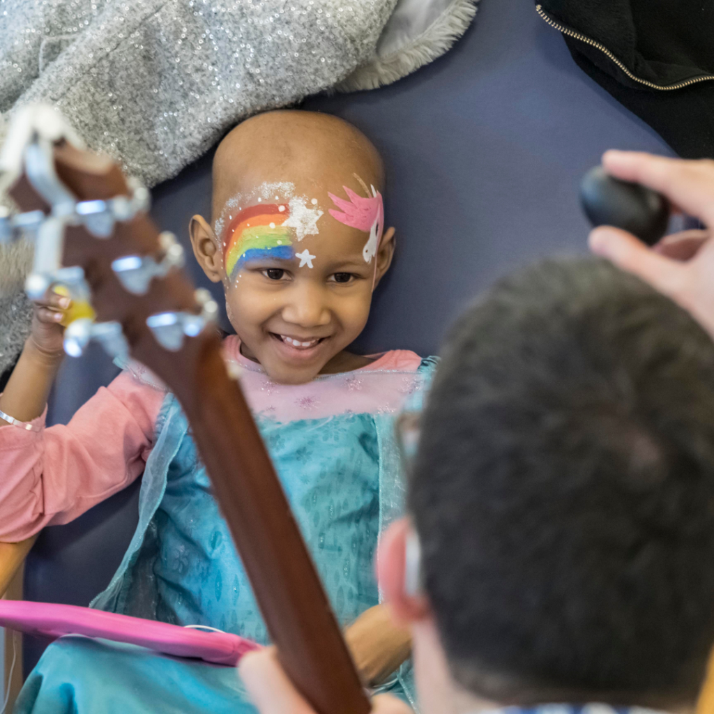 A photo of a little girl with a rainbow facepainted on her face making music with Nick